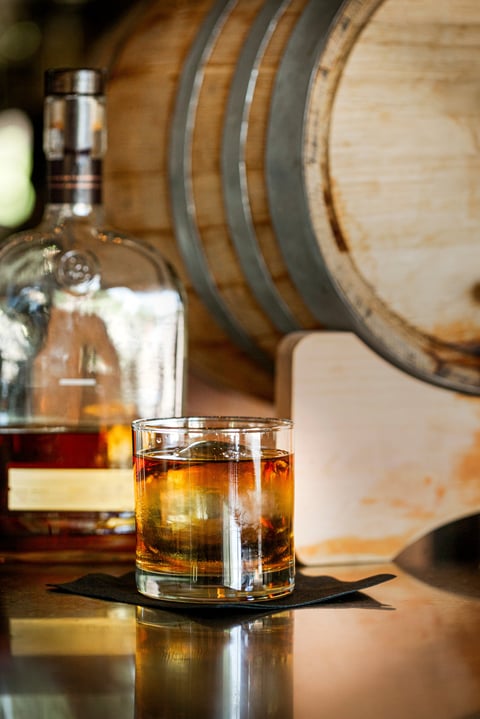 A glass of whiskey on a bar with a bottle and wooden barrel behind it