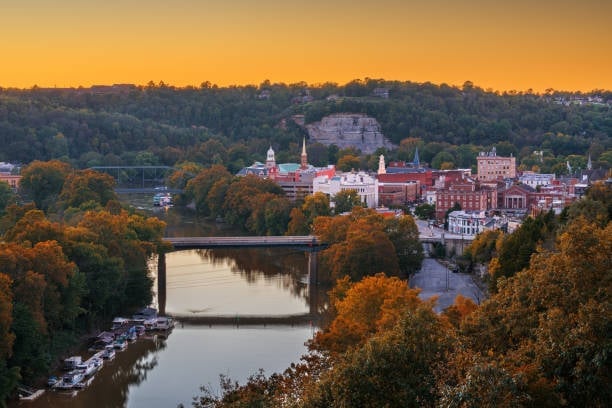 Small town cityscape at sunset with church steeple overlooking a river, surrounded by autumn foliage and wooded hills
